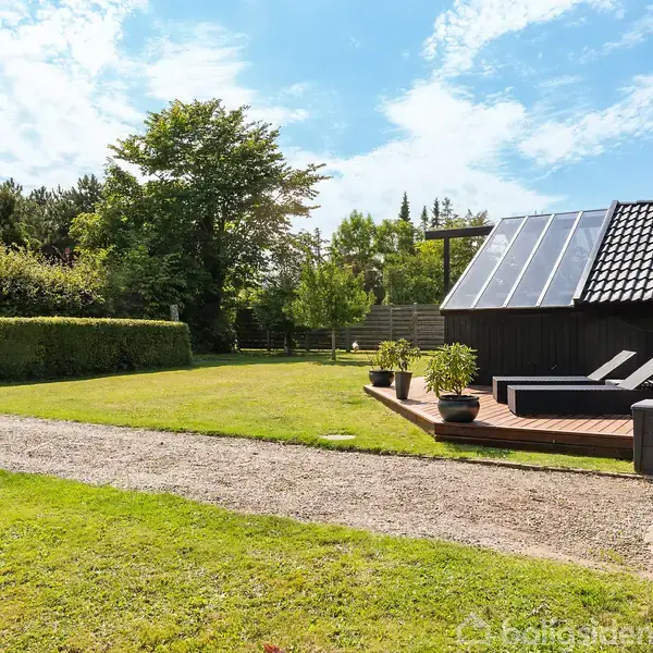 Grøn græsplæne foran et sortmalet hus med solpaneler, omgivet af træer og buske. Solskinsdag med blå himmel og få skyer. En sti løber langs græsplænen.