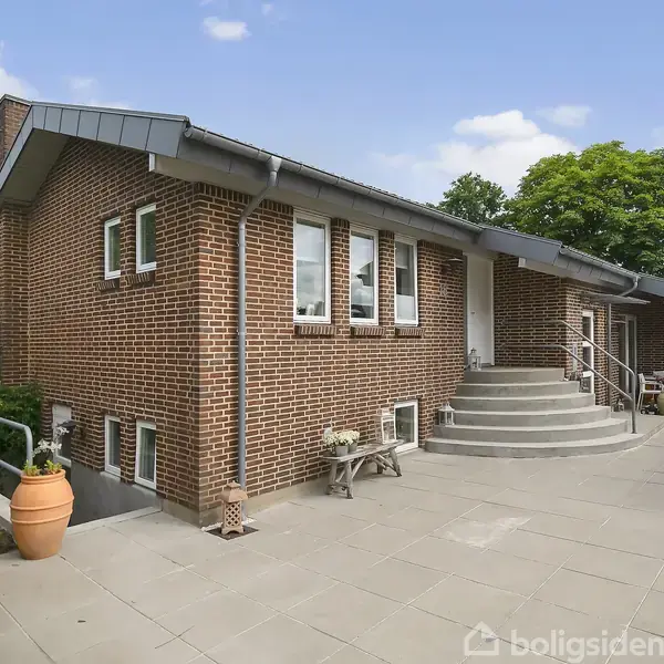 En murstensvilla med trappe fører op til hoveddøren. Villaen omgives af flisebelagt terrasse, grønne planter og en blå himmel.