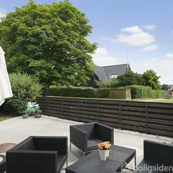 Terrassemøbler er arrangeret omkring et lille bord med blomster, mens en parasol er slået op. Omgivet af en have med grønne træer og et plankeværk, ses et hus i baggrunden.