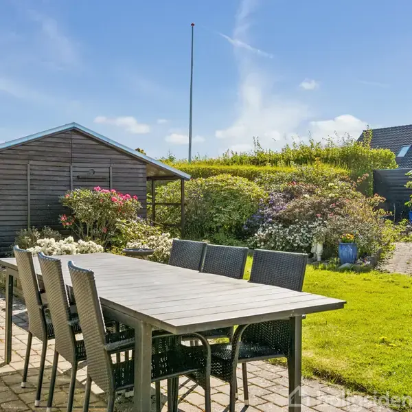 Havebord og stole står på en terrasse med fliser. I baggrunden ses en træskur omgivet af buske og blomster i en solrig have.