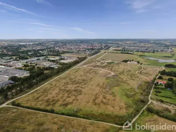Luftfoto af åbent grønt område med stier, omgivet af byens bebyggelse i det fjerne. Himlen er blå med få skyer, og området er let kuperet.