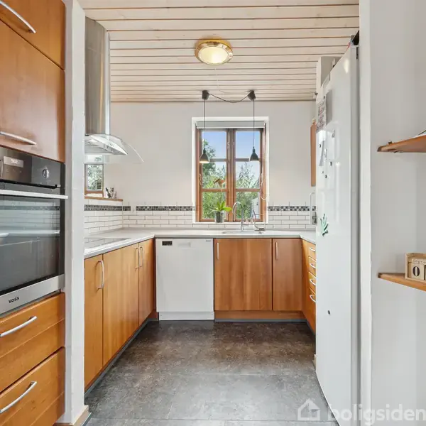 Et moderne køkken med træskabe og en indbygget ovn. En opvaskemaskine står ved siden af et vindue med udsigt til en grøn have. Flasker står på en hylde.