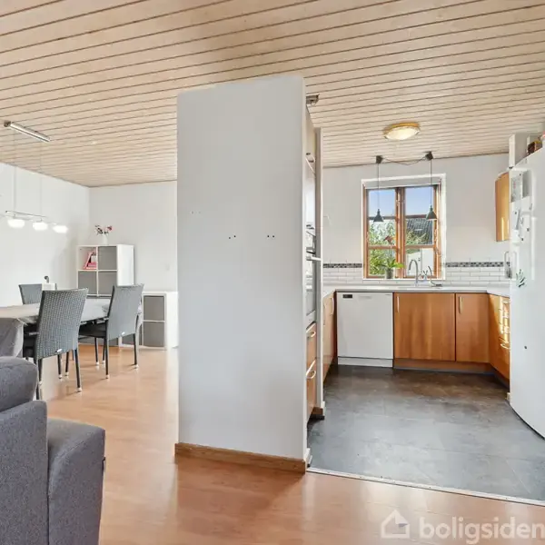 En stue og køkken i åben planløsning, hvor stuen indeholder spisebord med stole og sofa, og køkkenet har hvidevarer og vindue; trægulve og trælofter i rummet.