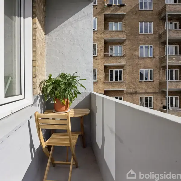 En balkon med et rundt, plant fyldt bord og en foldestol. Set i kontekst af en gråmalet mur med vinduer og en udsigt over en murstensbygning med altaner.