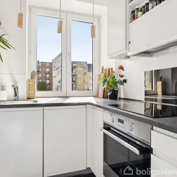 Et lyst køkken med hvide skabe og sorte bordplader. To store vinduer giver udsigt til bygninger udenfor. Hvidevarer inkluderer en ovn og kogeplade. Hylder til krydderier er over kogepladen.