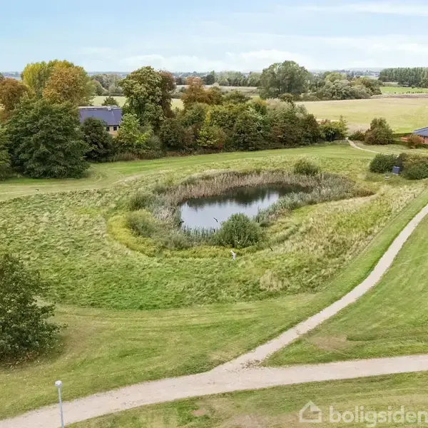 En lille sø omgivet af græsområde og træer. Stier snor sig gennem området, og der ses huse i baggrunden. Det er en åben, grøn parkagtig landskab.
