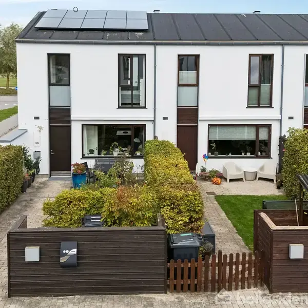 Rækkehus med solpaneler på taget, beliggende i et boligområde. Foran er der en have med hegn og buske, og en terrasse med stole og borde i baggrunden.