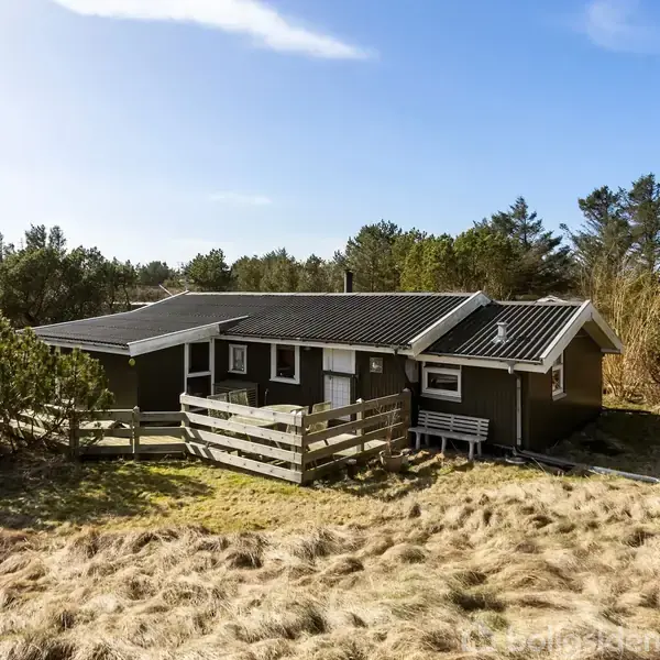 Et sommerhus med træbeklædning står omgivet af sandklitter og buske, under en blå himmel. En træterrasse med hegn ses foran huset. Ingen tekst er synlig.