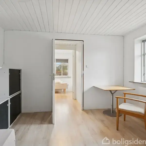 Et lyst rum med en åben dør, der fører til et andet værelse. To stole og et lille bord ved vinduet. Reol i hjørnet. Trægulv og hvidt loft.