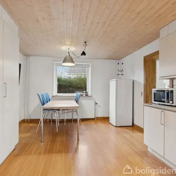 Et lyst køkken med træloft og trægulv. Der står et bord med blå stole i midten. Køkkenet har et vindue, et køleskab og skabe med hvide låger.