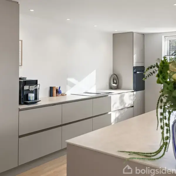 Et moderne køkken med grå kabinetter, en kaffemaskine, og indbygget ovn. En hvid bordplade prydes af en blåstribet vase med blomster. Store vinduer giver naturligt lys.