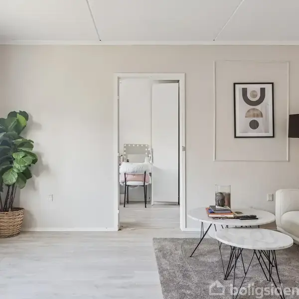 En stue med en lys sofa, sofabord og gulvlampe. Der hænger to abstrakte billeder på væggen. Til venstre står en stor plante ved siden af et vindue.