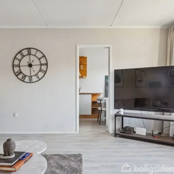 Stue med stort TV på stående bord. Rundt vægur hænger på væggen. Sofa og sofabord med bøger og dekorationer. Store vinduer med udsigt til træer udenfor.