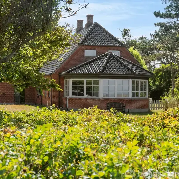 Et rødt murstenshus med sorte teglstenstagrender står stille omgivet af grøn vegetation og træer i en have. Solskinsvejr kaster lys og skygger over scenen.