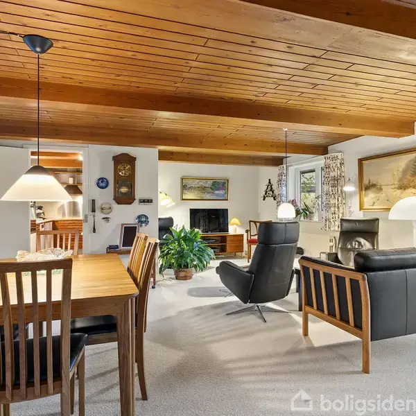 Et spisebord med stole står i en stue med åbent rum. Sofagruppe på højre side, pejs i baggrunden. Trælofter og hængende lamper skaber en hyggelig atmosfære.