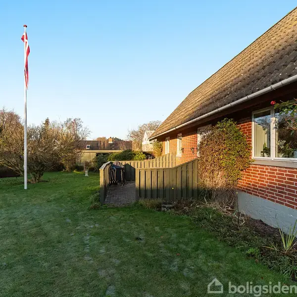 Et rødt murstenshus med vindue, omgivet af en velplejet grøn græsplæne. En flagstang med dansk flag står til venstre. Haven er kantet af buske og planter.