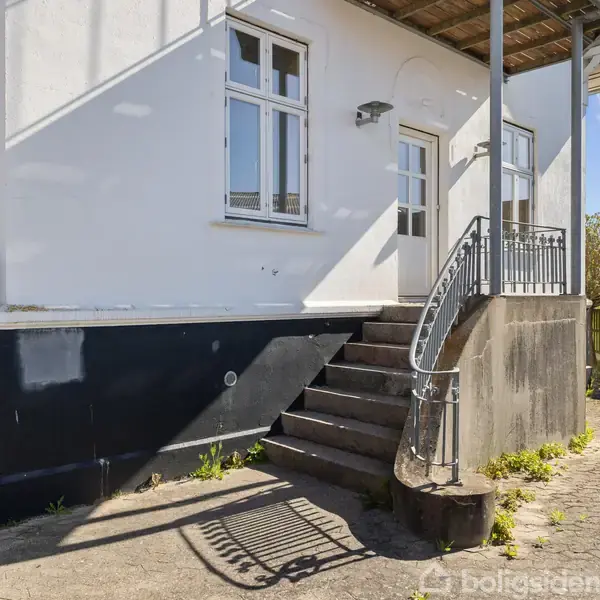Huset har en udvendig trappe, der fører op til en hvid dør. Trappen har et metalgelænder. Omgivet af flisebelagt gård og hegn. Solrig dag med blå himmel.