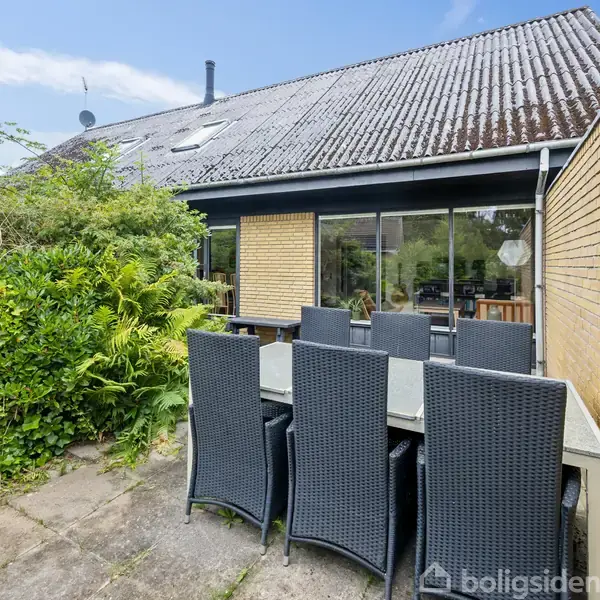 Udendørs terrasse med et sort spisebord og otte stole i naturligt lys. Omkranset af murstensvægge og grøn beplantning foran et hus med store vinduer og et skråt tag.