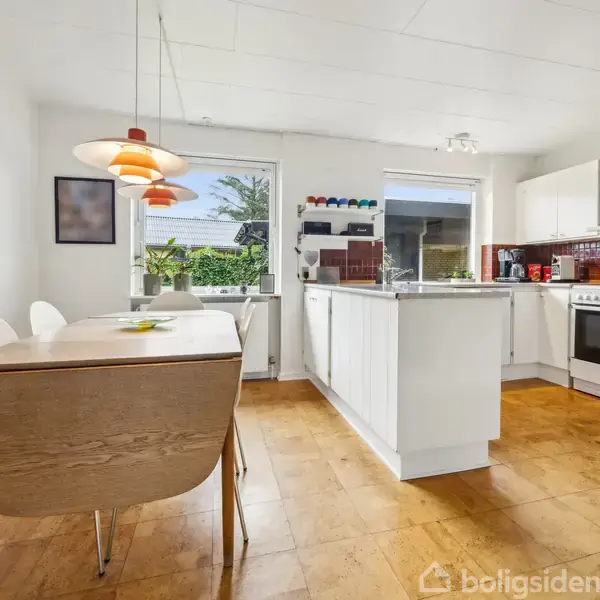 Køkken med hvidt interiør, spisebord, og pendellampe, aktion er ingen aktivitet. Kontekst er rummets lyse trægulv, store vinduer og udsigt til en grøn have.