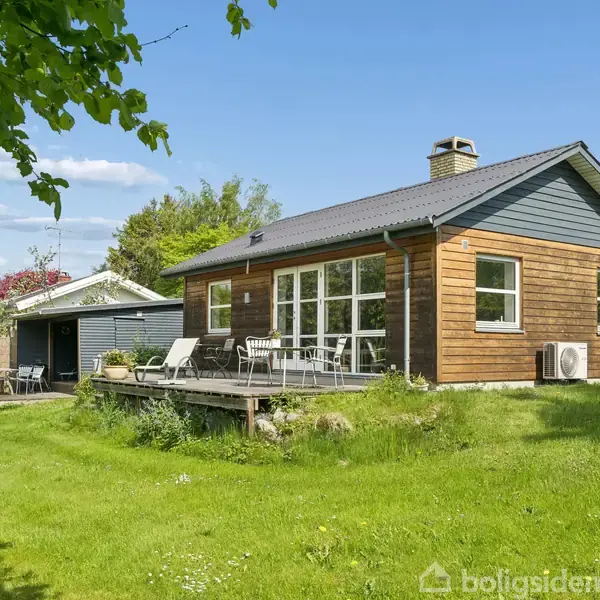 Stue med brændeovn står ved siden af et spisebord og stole. Sofaen ved vinduet har udsigt til terrassen. Lys strømmer ind gennem store vinduer.