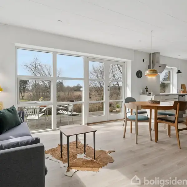 Stue med sofa og sofabord, åbner til køkken-alrum med spisebord og stole. Store vinduer giver udsigt til en have med træer. Lys og rummelig atmosfære.