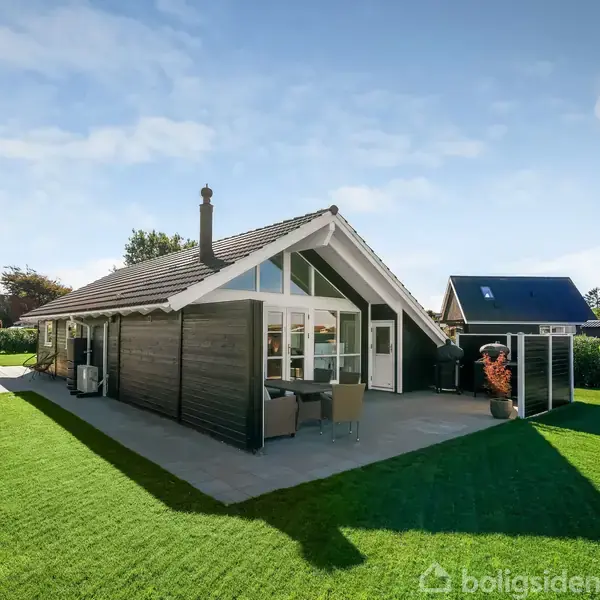 Et sortmalet sommerhus med store vinduer står på en velholdt græsplæne. Der er en terrasse foran huset med bord og stole. Omgivelserne er grønne og solbeskinnede.