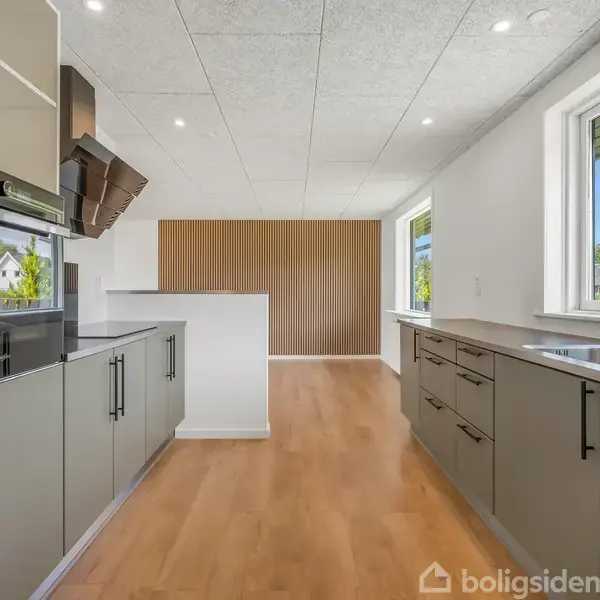 Køkken med moderne skabsmoduler, vask og komfur integreret. Rummelige træplankegulve strækker sig over rummet. Store vinduer giver udsigt til en frodig have omgivet af træer.