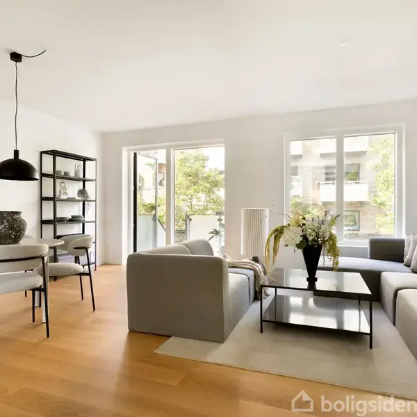 Stue med to grå sofaer arrangeret omkring et sofabord med blomsterbuket. Spisebord med fire stole og pendellampe. Store vinduer giver udsigt til grønne træer.