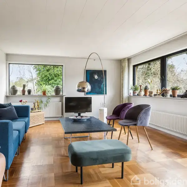En stue med en blå sofa, et lille tv på et bord, og store vinduer med udsigt til have. Stueplanter dekorerer vindueskarmen, og parketgulvet skaber en varm atmosfære.