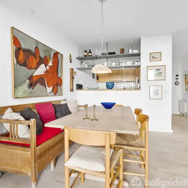 Langt, træbord med stole i en lys spisestue. Væggene er pyntet med moderne kunst. En lampeskærm hænger over bordet. I baggrunden ses en åben reol og forskellige dekorationer.