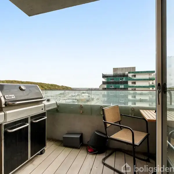 Grill står på en balkon med glasrækværk, ved siden af to stole og et bord, omgivet af moderne bygninger og udsigt over landskab.