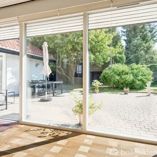 Glasskydedør åbner mod en solrig gårdhave med fliser. Havemøbler og en parasol står til venstre. En frodig busk skaber en grøn baggrund, omgivet af træer.