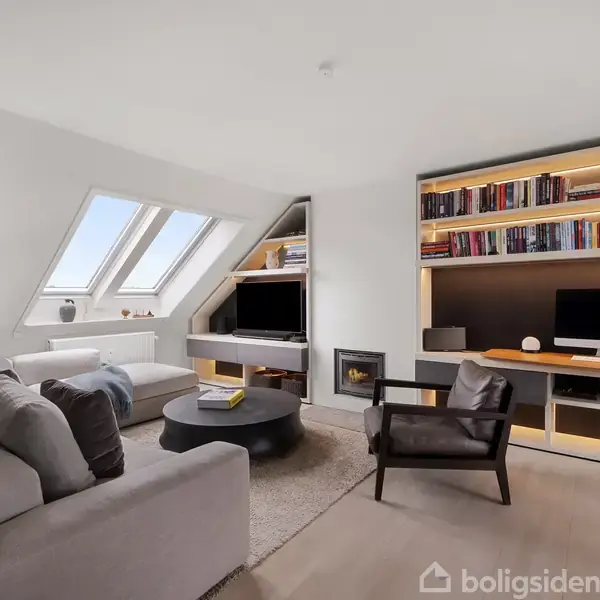 En moderne stue har en stor grå sofa, et rundt sofabord, og en reol med bøger. En computer står på et skrivebord. Skråvinduer lukker naturligt lys ind.