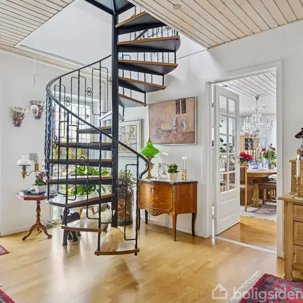 En sort snoet trappe forbinder to etager i en lys stue med trægulve. Møbler som kommoder og stole står omkring, og grønne planter pynter i det elegante rum.