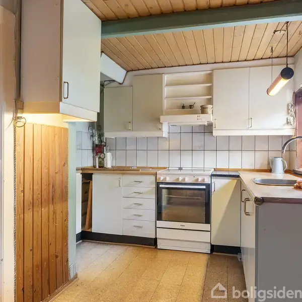 Et køkken med hvidt interiør og træloft. En komfur står centralt mellem skabe. Til højre, en vask ved et vindue. Vægur hænger på væggen. Belysning over køkkenbordet.