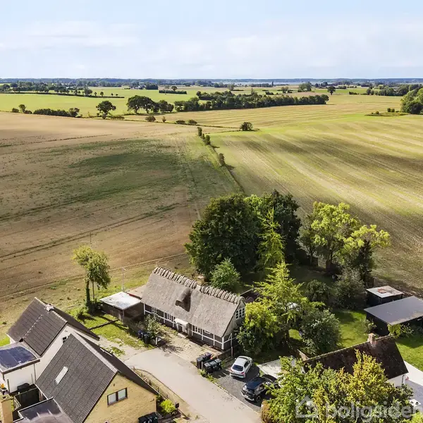 En landejendom omgivet af åbne marker, beplantede områder og en lang vejstrækning. Bygningerne, herunder huse og en carport med biler, ligger i et naturskønt og landligt område.