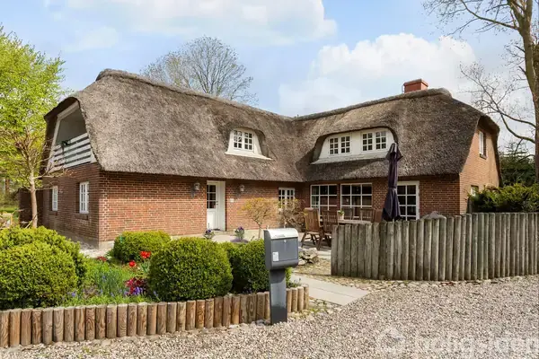 Stråtækt hus står stille omgivet af en grussti og velplejet have med buske og blomster. Terrasse med havemøbler og parasol til højre, indhegnet af lavt hegn.