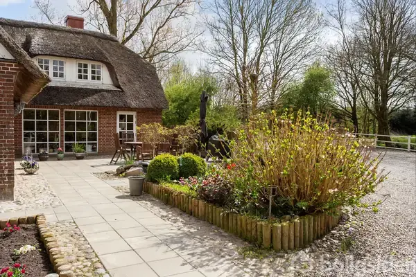 Stråtækt hus står stille ved siden af en flisebelagt terrasse. Omkring er en velplejet have med buske og blomster, og bagved ses træer, der skaber en fredelig og naturlig atmosfære.