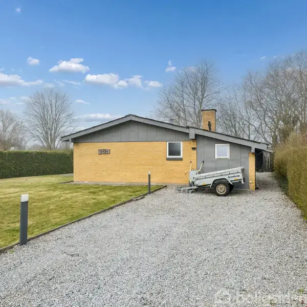 Et gult murstenshus med en trailer parkeret på grusvej foran. Omkranset af en velplejet have og hække, ligger huset i et beboelsesområde under en blå himmel.