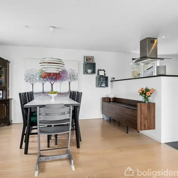Spisebord med seks stole står på et lyst trægulv i et moderne køkken-alrum. Over bordet hænger en rund lampe. I baggrunden er der et vitrineskab og blomsterdekoration.