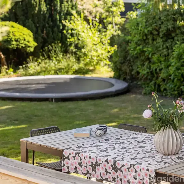 Et havebord med blomsterdug står på en terrasse foran en nedgravet trampolin og en frodig have. På bordet ligger en bog, en kop og en vase med blomster.