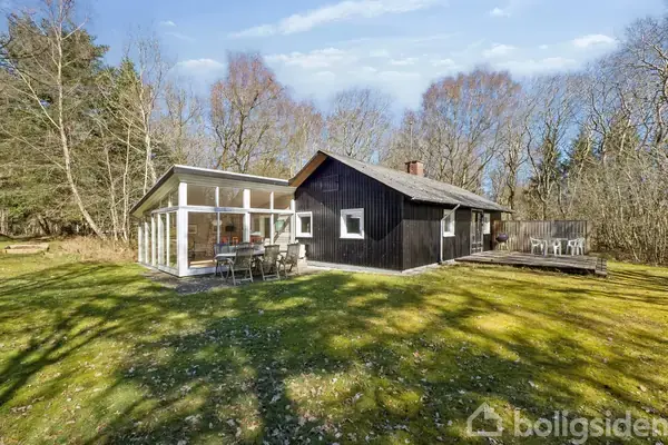 Køkkenet har hvide skabe og en emhætte over kogepladen. En dør fører til vaskerum med vaskemaskine. Vinduet viser træer udenfor, omgivet af lyse lamper og loft.