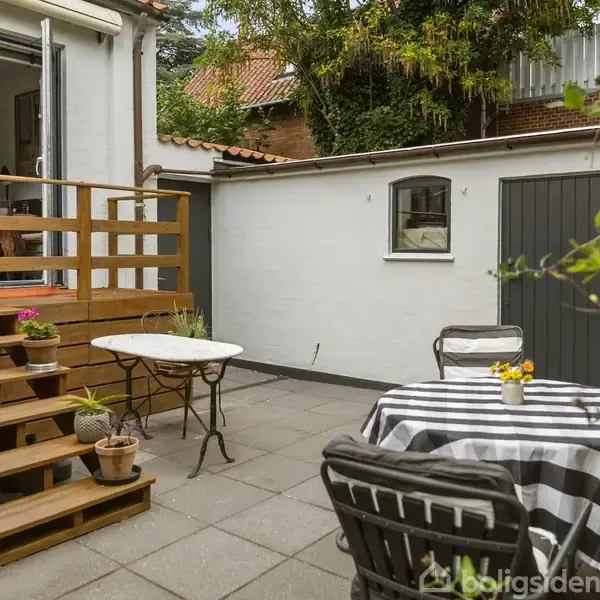 Et bord med sort-hvid-stribet dug står på en flisebelagt terrasse, omgivet af stole. Trætrapper fører ind til et hus, og planter dekorerer området.