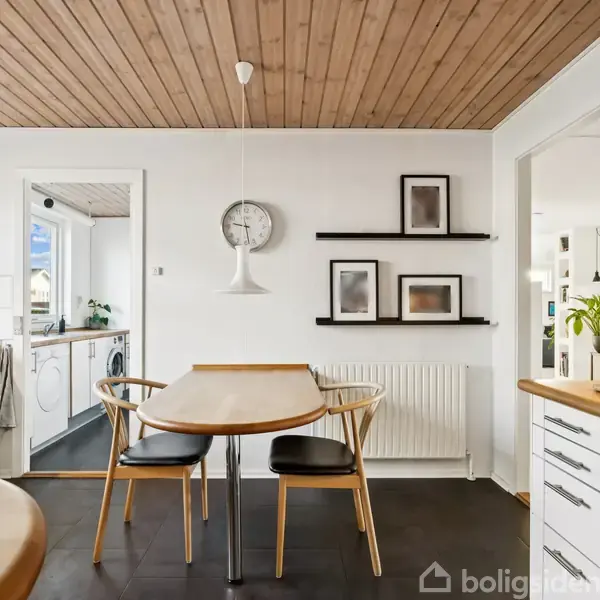 Træbord med fire stole står i et lyst køkken med hvide skabe og sorte fliser. Ur og billeder på væggen. Dør til bryggers med vaskemaskine ses.