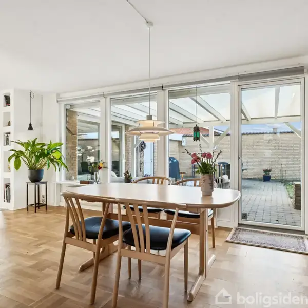 Et spisebord med fire stole står foran en stor glasvæg med udsigt til en terrasse. Der hænger en lampe over bordet, og en plante står ved siden af.