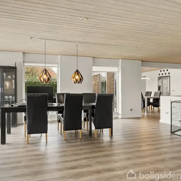 Spisebord med sorte stole står i et lyst rum med trægulv og store vinduer, der giver udsigt til en have. En brændeovn ved siden af giver varme og hygge.