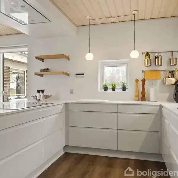 Et moderne køkken med hvide skuffer og åbne hylder. To hængelamper lyser rummet, mens en kaffemaskine og køkkenredskaber står på bordpladen. Store vinduer og træloft skaber lys atmosfære.