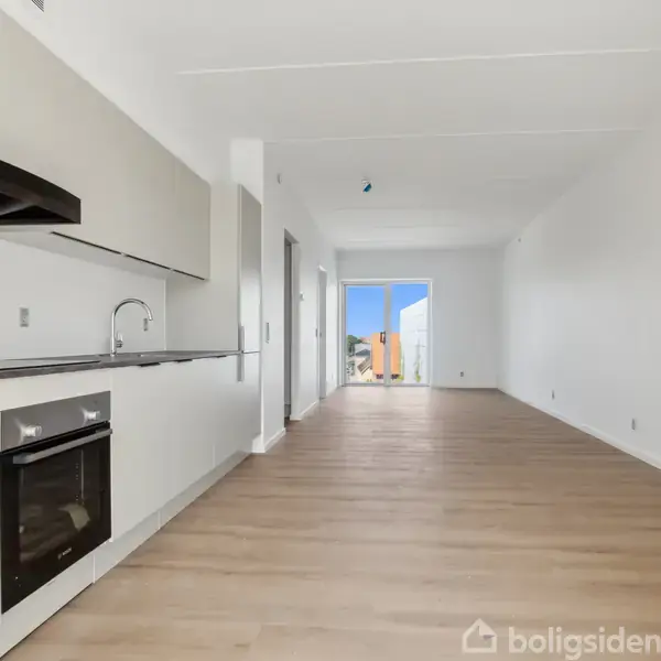 Et moderne køkken med hvide skabe og indbygget ovn strækker sig langs en væg. Trægulve fører til en lys stue med dør til balkon.