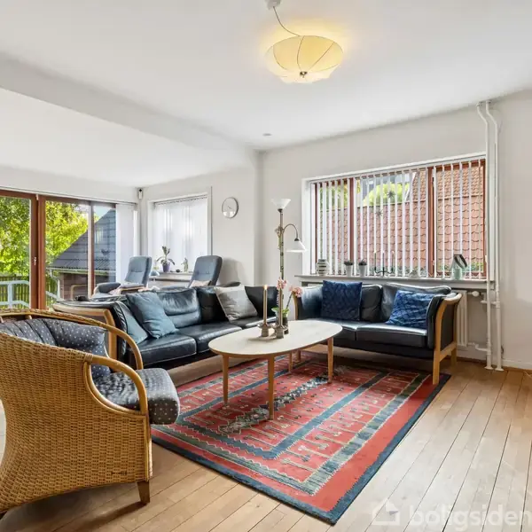 En stue med sorte sofaer og et rundt bord står på et orientalsk tæppe. Store vinduer slipper lys ind, og udenfor ses et grønt træ. En lampe hænger i loftet.