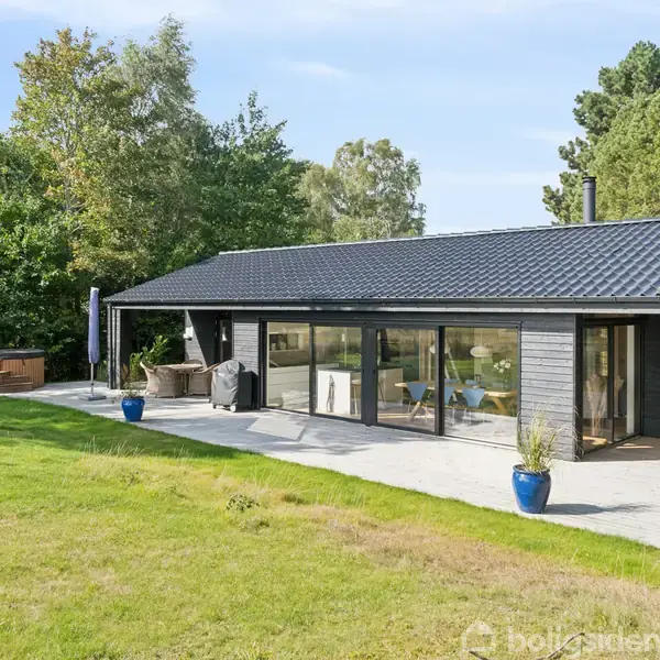 En moderne bungalow med sorte træpaneler og store vinduer står på en terrasse. En græsplæne og træer omkranser huset. Blå blomsterpotter pynter terrassen i den solrige have.